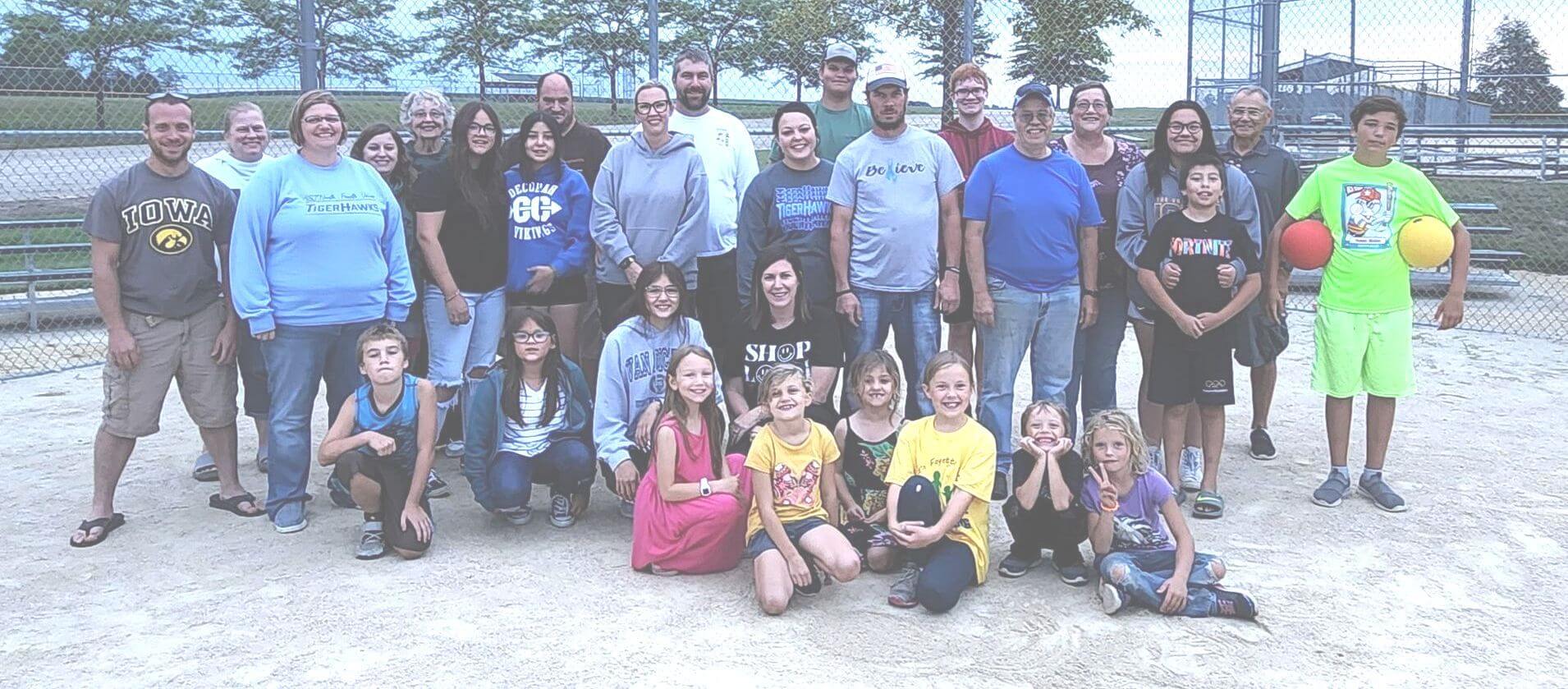 A group of adults and children pose together on a baseball field, smiling at the camera.
