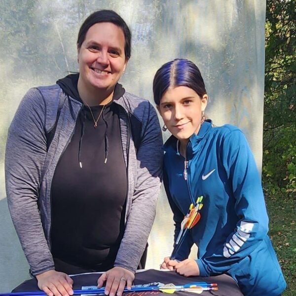 Two people smile outdoors by a table with arrows and archery gear, trees in the background.