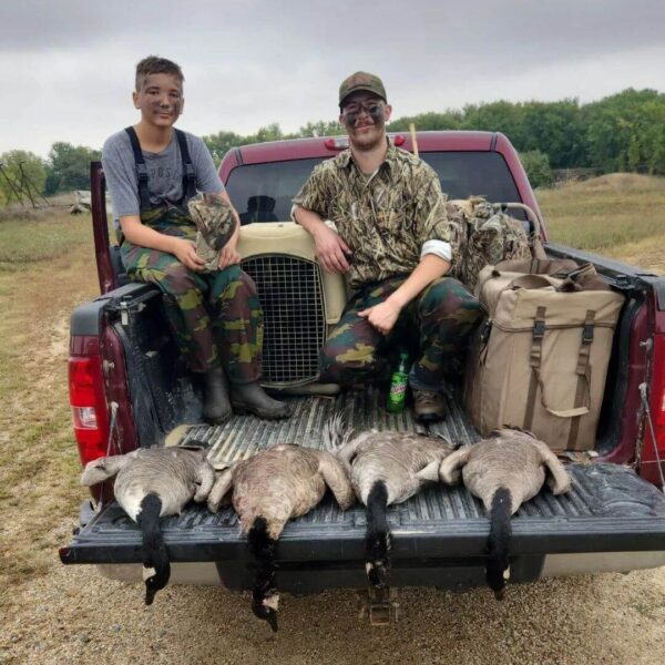 Two people in camo sit on a truck bed with hunted geese laid out, smiling after a successful hunt.