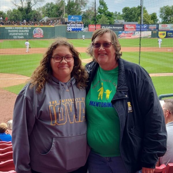 Two women smiling at a baseball stadium, standing in front of red seats with the field in the background.