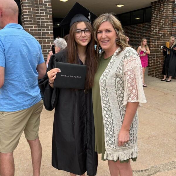 Graduate in cap and gown holding diploma, standing with a woman, both smiling outdoors after ceremony.