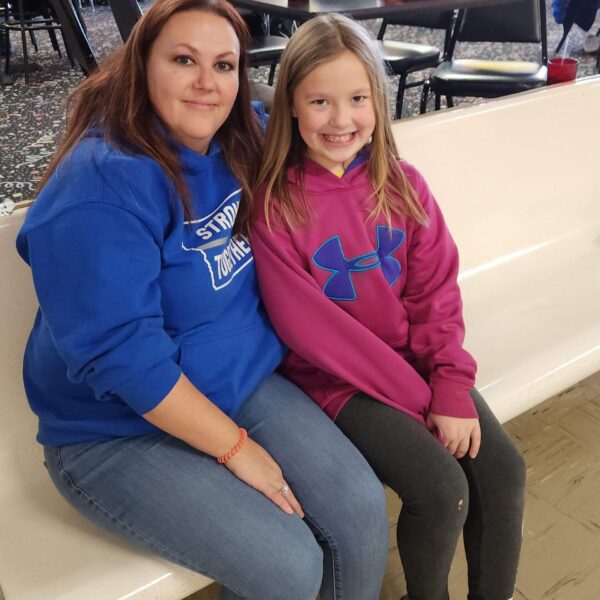 A woman and a girl sit smiling on a bench at a bowling alley, both wearing hoodies.