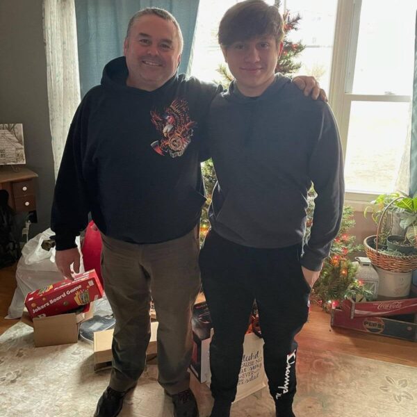 Two people stand smiling by a decorated Christmas tree in a living room with gifts on the floor.