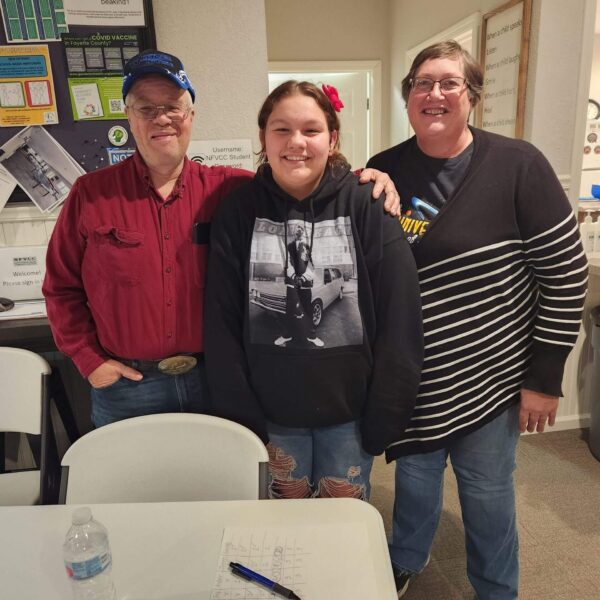 Three people smile together indoors near a table with a water bottle, pen, paper, and a Phase 10 card game.