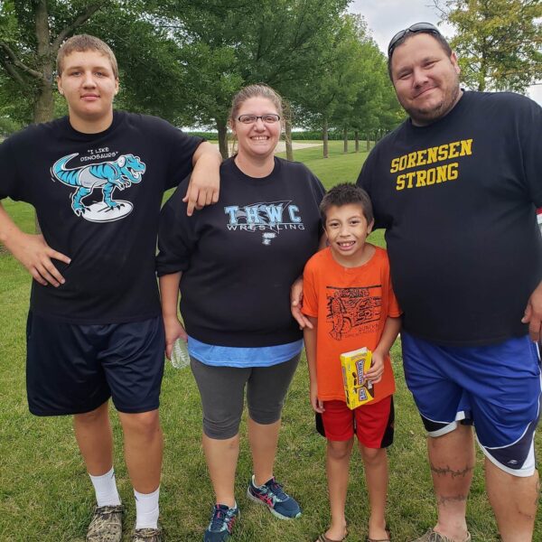 Four people stand smiling outdoors on grass, with trees in the background, on a cloudy day.