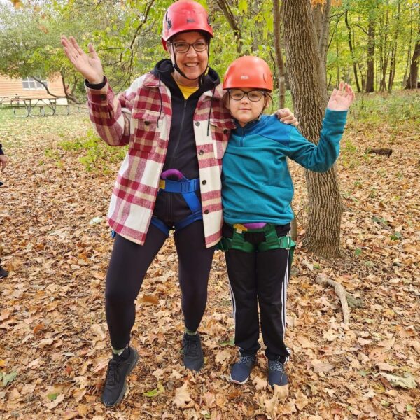 Two people in helmets and harnesses smile and pose outdoors in a forest with autumn leaves on the ground.