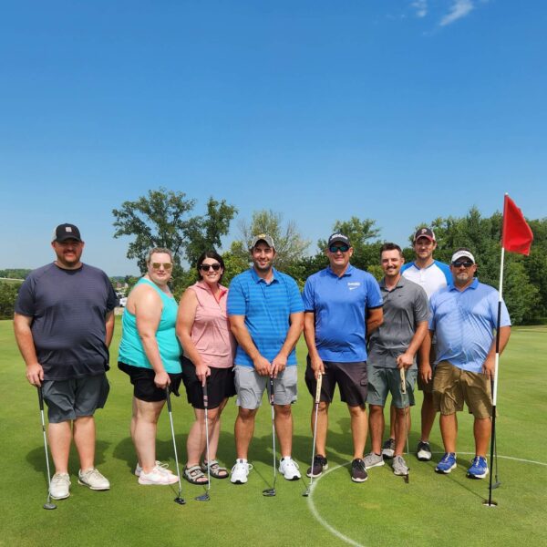 Eight people stand together on a golf course near a red flag, posing with golf clubs on a sunny day.