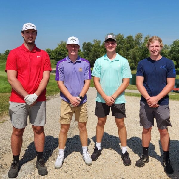 Four young men stand side by side outdoors on a golf course, smiling in casual golf attire.