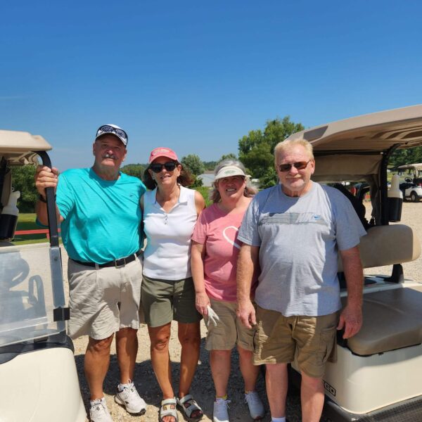 Four adults in casual summer clothes stand smiling between two golf carts on a sunny day.