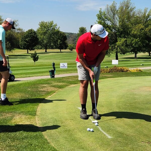 A man in a red shirt putts on a golf green while another man watches nearby on a sunny day.