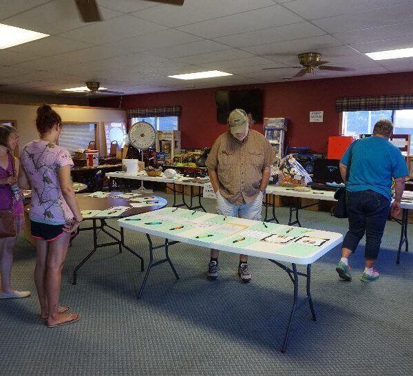 Four people look at items and papers displayed on tables in a community room with various objects in the background.