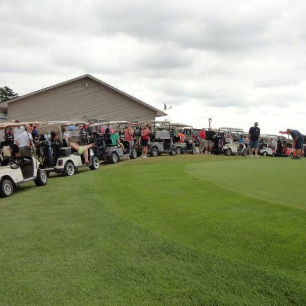 Golfers stand by golf carts lined up near a putting green on a cloudy day at a golf course.