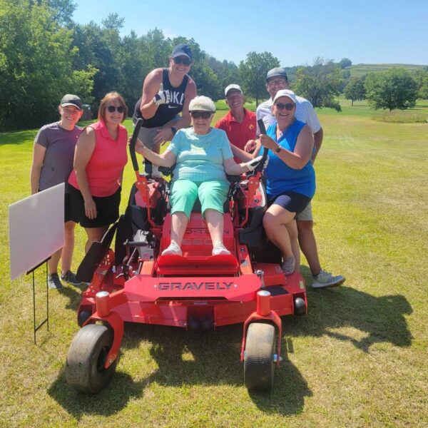 Seven smiling people pose around a large red lawn mower on a sunny day in a grassy, tree-filled park.