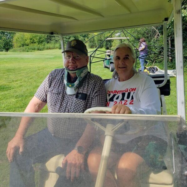 An older man and woman sit together in a golf cart on a sunny day, smiling at the camera.