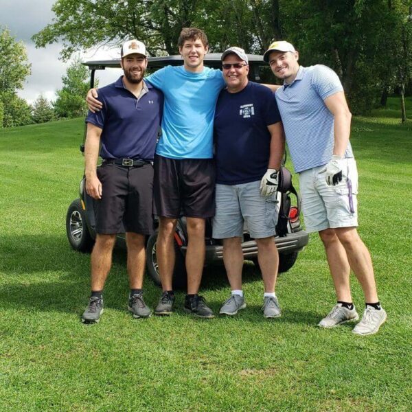 Four men stand smiling together on a golf course in front of a golf cart, surrounded by green trees and grass.