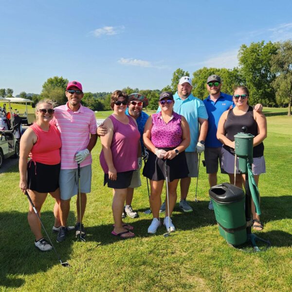 A group of eight adults with golf clubs pose together on a sunny golf course.