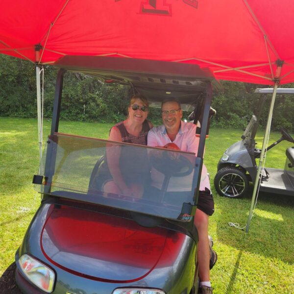 Two people smiling and sitting together in a golf cart under a red tent with "State" printed on it.