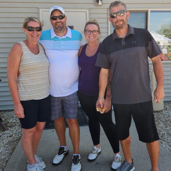 Four adults smiling and standing together outside a building on a sunny day.