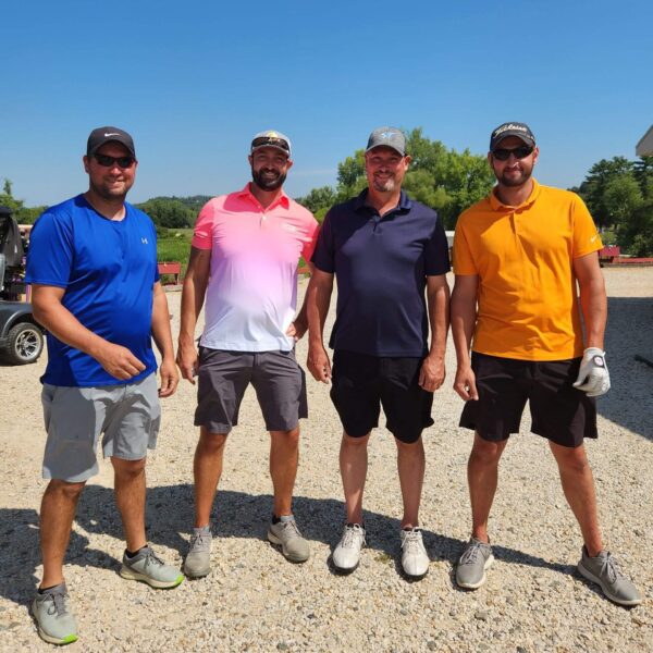 Four men in colorful shirts and shorts stand together outdoors on a sunny day, posing for a group photo.