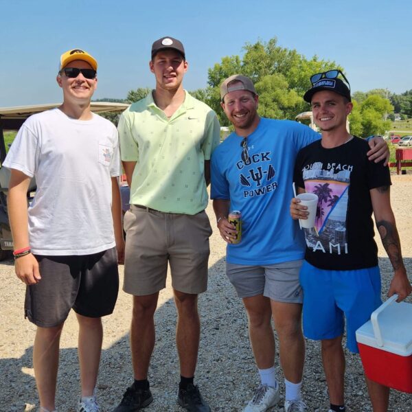 Four men stand outdoors on gravel, smiling at the camera; two hold drinks, one holds a cooler.