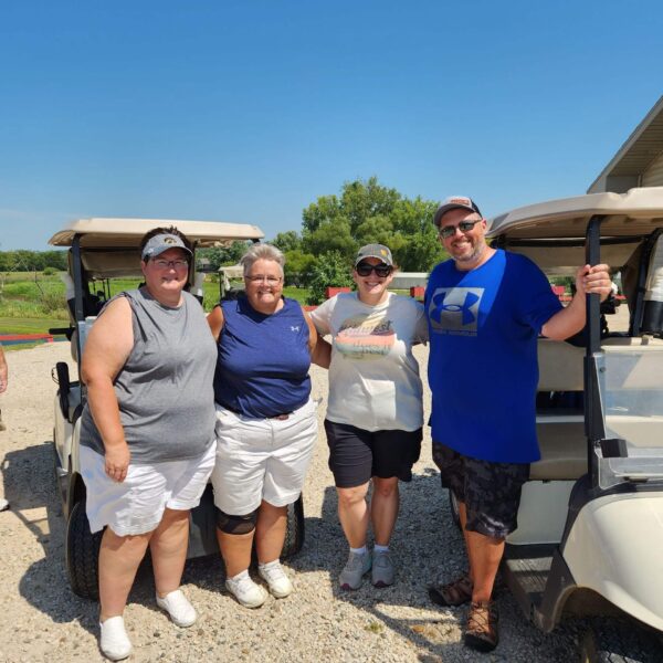 Four adults stand smiling together by golf carts on a sunny day, dressed in casual summer clothes.