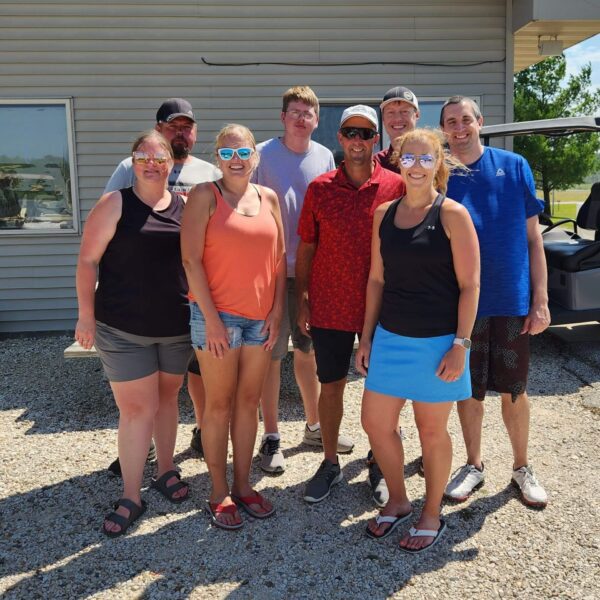 Eight smiling adults pose together outside on a sunny day, standing on gravel in front of a building.