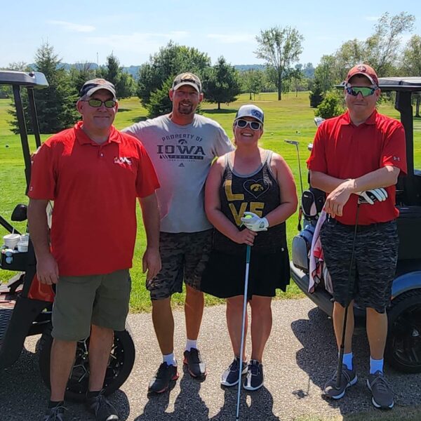 Four people pose on a sunny golf course, standing by two golf carts with clubs, wearing casual golf attire.