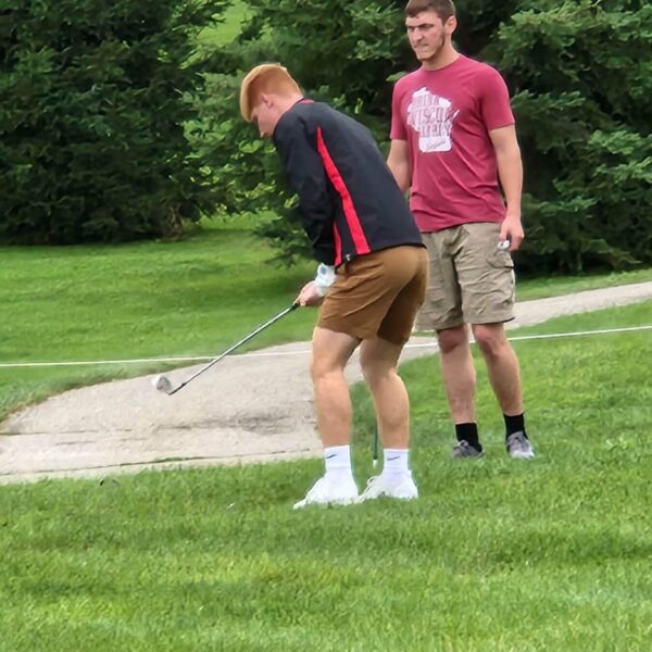Two young men on grass; one is swinging a golf club while the other stands watching in casual clothes.