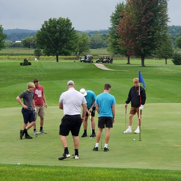 Six men stand on a golf green, some looking at the hole, with trees and golf carts in the background.