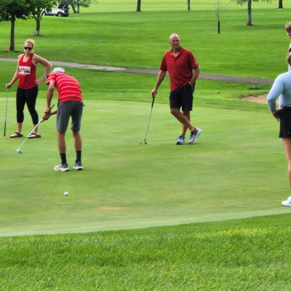 Four people watch as a man in a striped shirt putts on a green golf course, with trees in the background.