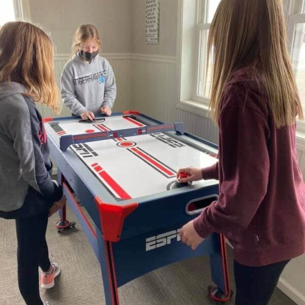 Three girls play air hockey together indoors, one wearing a mask, with windows in the background.