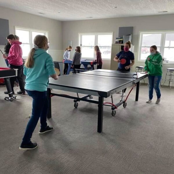 Several teenagers play ping pong and foosball in a bright room with large windows and gray carpeting.