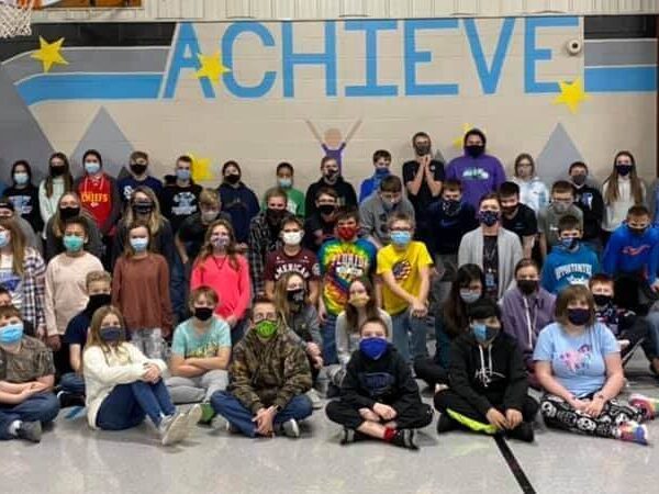 A large group of masked children and adults pose together in a gym with "ACHIEVE" painted on the wall.