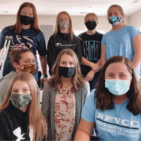 Eight girls wearing face masks pose together indoors, some standing and some sitting, smiling at the camera.