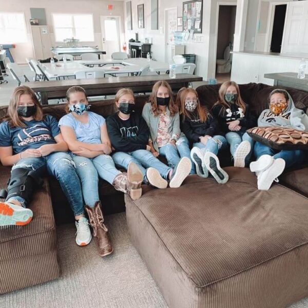 Seven girls wearing face masks sit closely together on a brown sectional couch in a bright room.