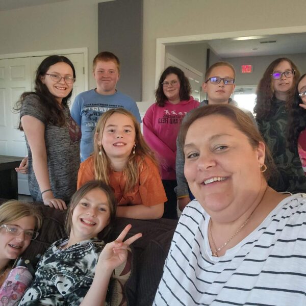 A woman and nine smiling kids pose for a cheerful group selfie in a living room.
